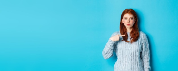 Foto gratuita ragazza rossa nervosa e confusa che punta a se stessa in piedi in maglione su sfondo blu