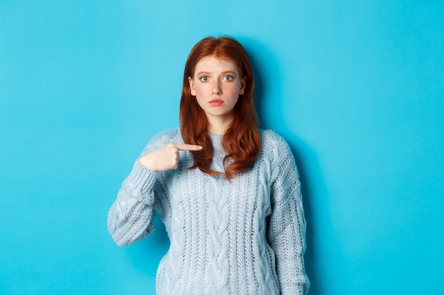 Nervous and confused redhead girl pointing at herself, standing in sweater against blue background.