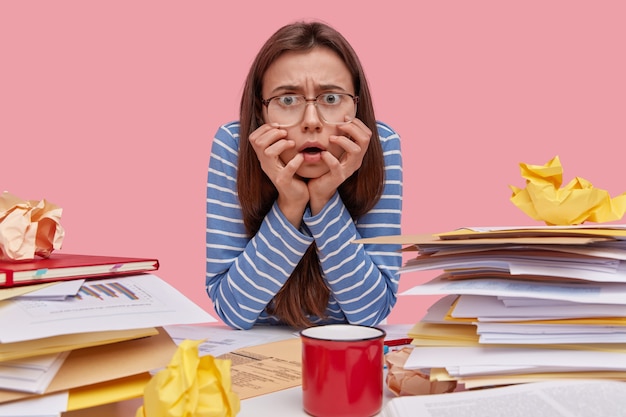 Nervous displeased lady stares directly at camera keeps hands under chin, wears blue striped jumper, square glasses, afraids of deadline