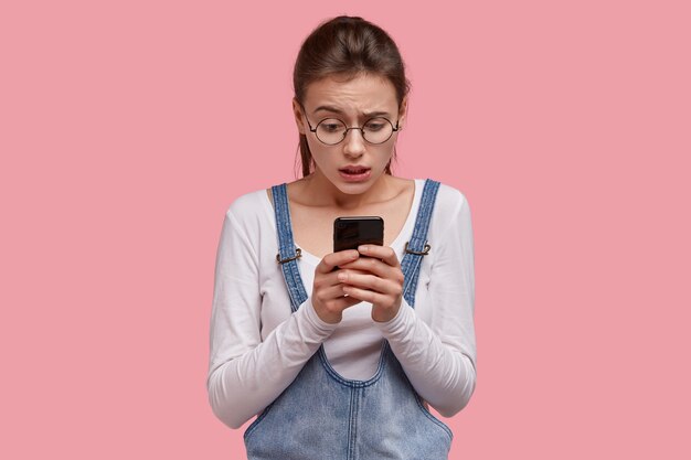 Nervous displeased woman holds modern cell phone, puzzled to recieve not much likes under her post in social networks, wears casual clothes