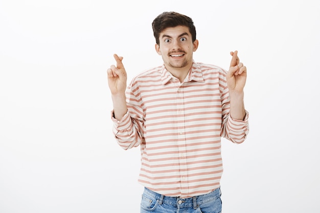 Foto gratuita ragazzo nervoso amichevole in attesa di risultati. ritratto di bell'uomo europeo gentile con barba e baffi, incrociando le dita, desiderando e pregando per il successo, in piedi con il sorriso sul muro grigio