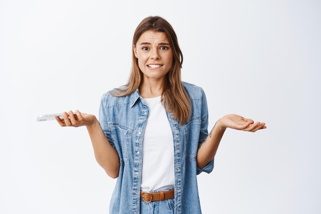 Foto gratuita la ragazza nervosa allarga la mano lateralmente complicata e morde il labbro, tiene in mano lo smartphone, non sa nulla, non capisce cosa fare, in piedi preoccupata sul bianco