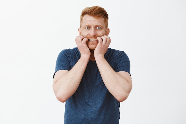 Nervous good-looking redhead guy with bristle, biting fingernails and staring anxious and shook, feeling scared over gray wall