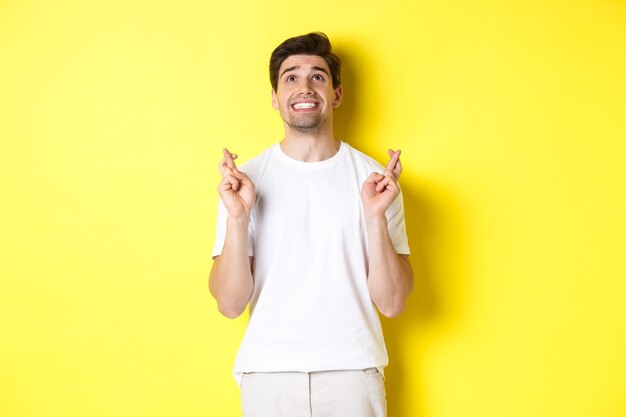 Nervous and hopeful man praying to god, making wish with fingers crossed, panicking and standing over yellow background