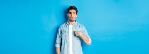 Free photo nervous man pointing at himself and looking confused standing in casual clothes over blue background