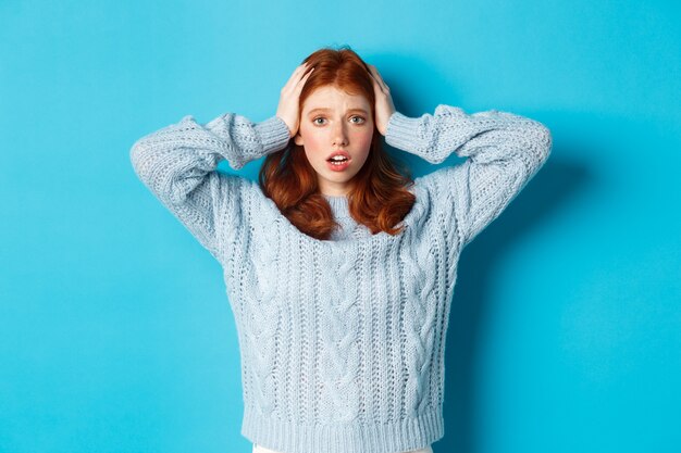 Nervous redhead girl standing overwhelmed, holding hands on head in panic and staring at camera, standing anxious against blue background.