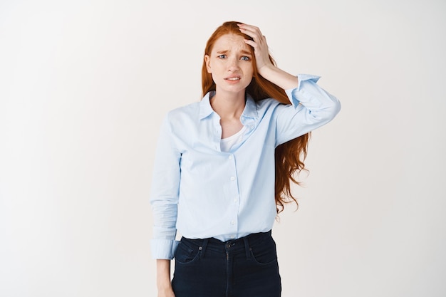 Free photo nervous redhead woman looking worried and upset, touching head and looking at at front guilty, forgot something, standing over white wall