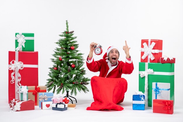 Nervous santa claus sitting on the ground and raising clock near gifts and decorated Xmas tree on white background
