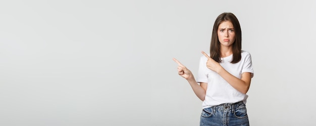 Foto gratuita ragazza carina timida nervosa che punta le dita nell'angolo in alto a sinistra e sembra triste avendo rimpianto