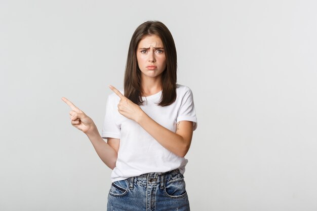 Nervous timid cute girl pointing fingers upper left corner and looking sad, having regret.