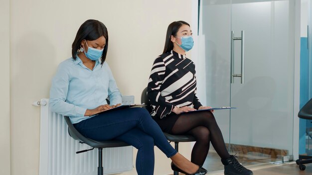 Nervous women waiting in queue to join job interview, preparing with cv resume for HR meeting. African american woman attending appointment with executive company manager during pandemic.