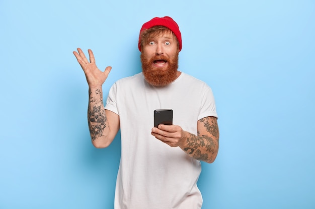 Nervous worried bearded ginger guy raises hand, holds mobile phone, wears red hat and white t shirt, uses modern technology, feels perpelexed and troubled, checks bill list online, gestures angrily