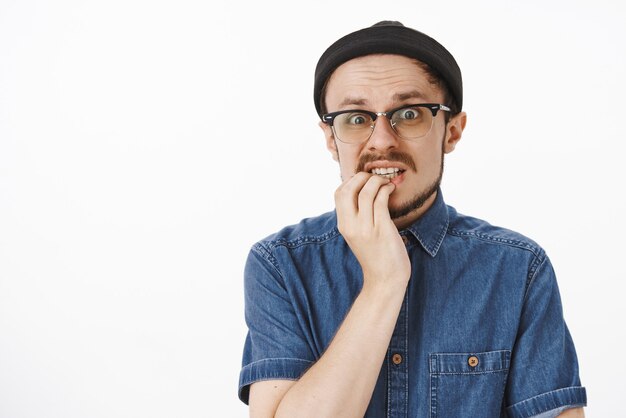 Nervous and worried concerned good-looking male with beard in glasses trendy beanie and shirt holding hand on lower lip staring shook and anxious making huge terrible mistake