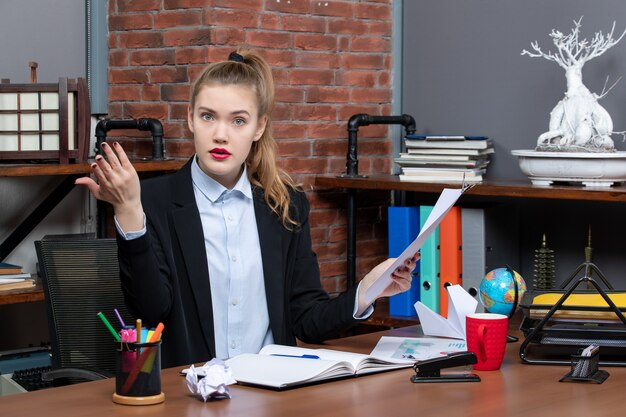 Nervous young lady sitting at a table and showing the document in the office