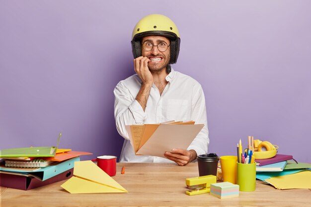 Nervous young male designer holds paper documents, bites finger nails, wears protective helmet and white shirt, sits at desktop with different things