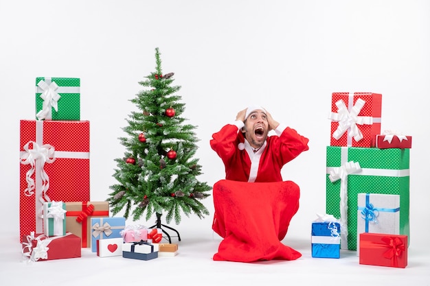 Free photo nervous young man dressed as santa claus with gifts and decorated christmas tree sitting on the ground on white background