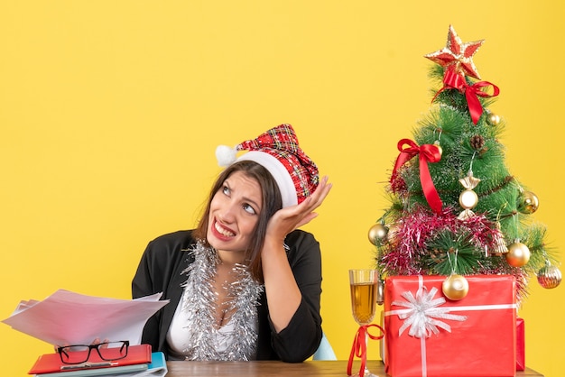 Donna d'affari nervosa in vestito con cappello di babbo natale e decorazioni di capodanno che tiene documenti e si siede a un tavolo con un albero di natale su di esso in ufficio