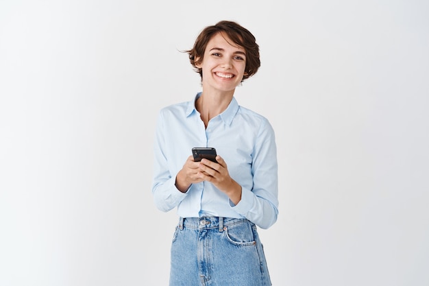 Foto gratuita rete. donna professionale sorridente che tiene smartphone in piedi sul muro bianco