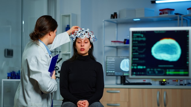 Neurological doctor cheking eeg headset explaining to patient diagnosis of disease and treatment for nervous system. Scientist researcher analysing brain scan and tomography on monitor in lab