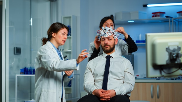 電気活動を分析する脳スキャンの準備をしている医学者が脳波スキャンヘッドセットを調整している間、モニターに向けて治療結果を説明する神経学研究者