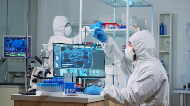 PPE 슈트를 입은 신경과 전문의는 PC에 장비를 갖춘 실험실에서 백신 개발 작업을 하고 있습니다. covid19에 대한 치료 개발 연구를 위해 첨단 기술을 사용하여 바이러스 진화를 조사하는 팀