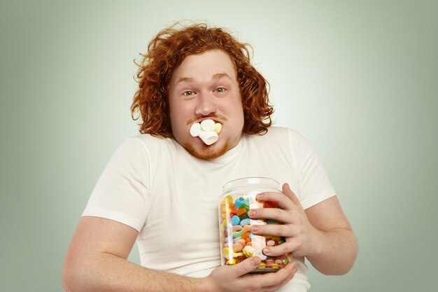 Never enough. Close up shot of funny young plump man with curly ginger hair holding tight jar of candies, looking with startled surprised expression, his mouth stuffed with marshmallow