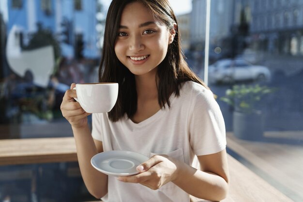 お気に入りのカフェに立ち寄って、おいしいコーヒーを飲むのを忘れないでください。短い黒髪のプレートとカップドリンクのカプチーノを持った魅力的なアジアの女の子が都会の空間で元気にリラックスして笑っています。