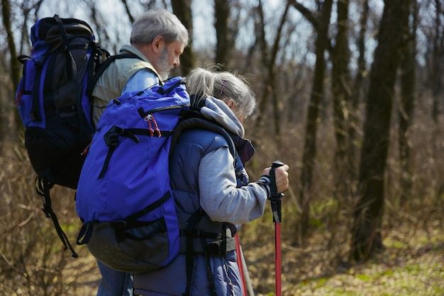 New cool experience. Aged family couple of man and woman in tourist outfit walking at green lawn near by trees in sunny day. Concept of tourism, healthy lifestyle, relaxation and togetherness.