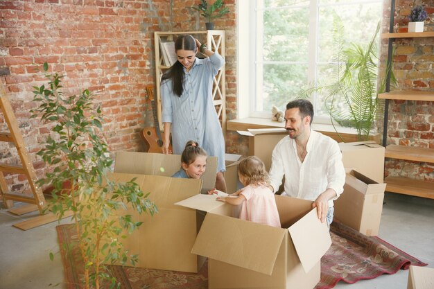 Новая жизнь. Взрослая семья переехала в новый дом или квартиру. Супруги и дети выглядят счастливыми и уверенными. Переезд, отношения, новая концепция жизни. Распаковываем коробки со своими вещами, вместе играем.