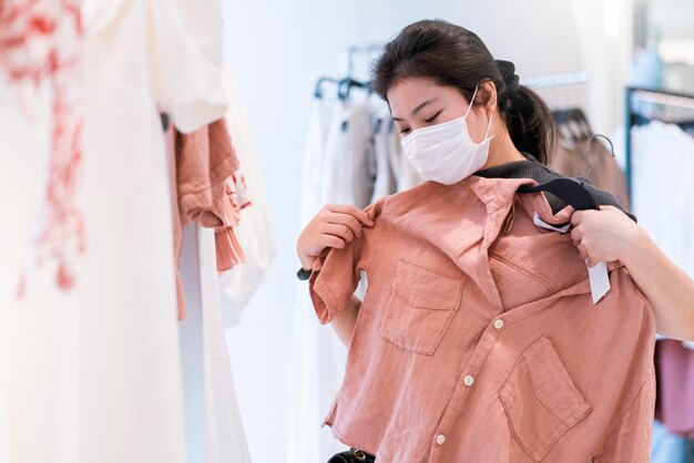 デパートモールの背景にあるブティックショップの新しいライフスタイルで、流行の若者アジア人女性がフェイスマスク保護ショッピングドレスや布を着用した後の新しい通常