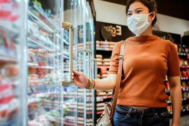 New normal after covid epidemic young smart asian female shopping new lifestyle in supermarket with face shild or mask protection hand choose fresh vegetable of fruit new normal lifestyle