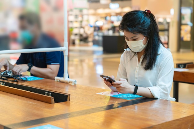 Новое нормальное социальное дистанцирование с азиатской женщиной в маске для лица защита от вирусов в ожидании сидеть в кресле использовать связь смартфона с космическим расстоянием для предотвращения распространения вируса короны