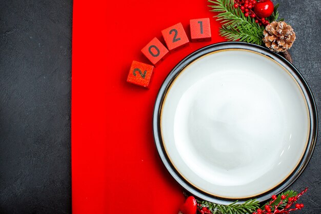 Новогодний фон с аксессуарами для украшения тарелки еловые ветки и числа на красной салфетке на черном столе горизонтальный вид