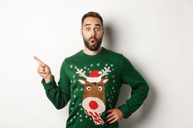 New year celebration and winter holidays concept. Surprised man in christmas sweater pointing finger at upper left corner, looking amazed, white background