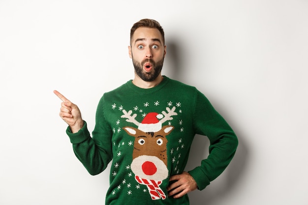 New year celebration and winter holidays concept. Surprised man in christmas sweater pointing finger at upper left corner, looking amazed, white background