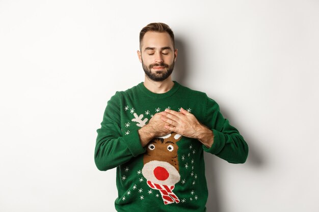 New year celebration and winter holidays concept. Young relaxed man feeling nostalgic, holding hands on heart and close eyes, dreaming about something on Christmas