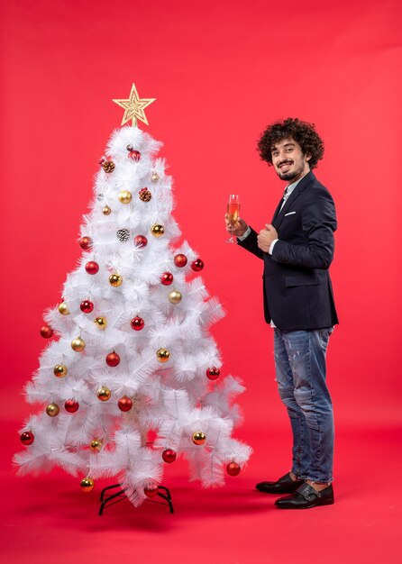 Празднование нового года с молодым человеком, держащим бокал вина возле украшенной белой елки на красном