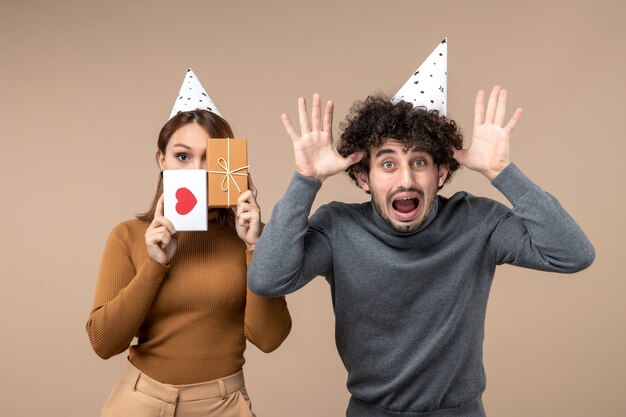 New year concept with young couple wear new year hat girl closing her face with heart and gift and guy showing ten on gray