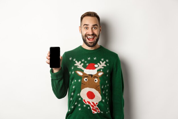 新年、祝日、お祝い。白い背景の上に立って、スマートフォンの画面を表示して、クリスマスセーターで興奮したひげを生やした男