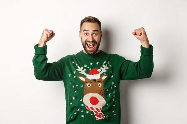 Новый год, праздники и праздник. Облегченный и взволнованный мужчина в рождественском свитере, поднимающий руки и празднует, торжествуя, стоя на белом фоне