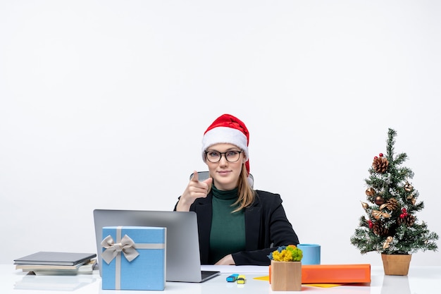 New year mood with blonde woman with a santa claus hat sitting at a table with a Christmas tree and a gift on it on white background