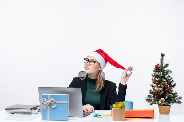 New year mood with dreamy positive blonde woman with a santa claus hat sitting at a table with a Christmas tree and a gift on it on white background