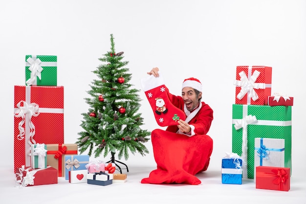 Free photo new year mood with funny positive santa claus sitting on the ground and showing christmas sock near gifts and decorated xsmas tree on white background