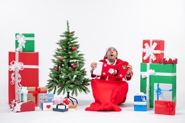 New year mood with funny positive surprised shocked santa claus sitting on the ground and showing christmas sock near gifts and decorated xsmas tree on white background