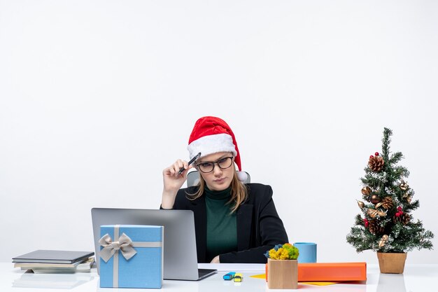 New year mood with indecisive blonde woman with a santa claus hat sitting at a table with a Christmas tree and a gift on it on white background