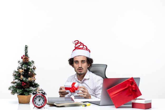 New year mood with sad unsatisfied young businessman with santa claus hat sitting in the office and holding his gift on white background