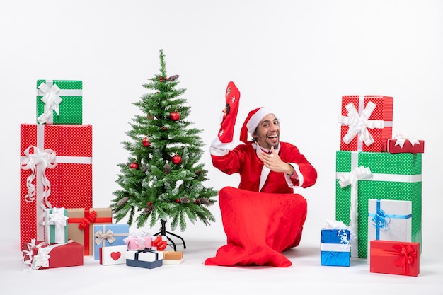 Umore di nuovo anno con babbo natale seduto per terra e indossare calzino di natale vicino a regali e albero di natale decorato su sfondo bianco stock photo