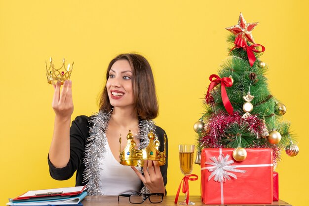 Новогоднее настроение с улыбающейся очаровательной дамой в костюме, держащей короны в офисе на желтом изолированном