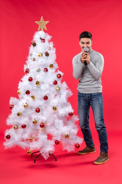 Новогоднее настроение с улыбающимся позитивным парнем в джинсах, стоящим возле украшенной елки на красном