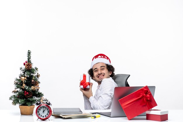New year mood with smiling positive young businessman with santa claus hat sitting in the office and pointing his gift happily on white background